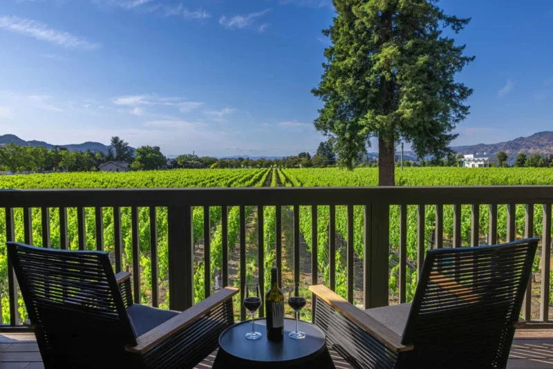Two outdoor chairs and wine on a balcony with a stunning view of green napa valley vineyard rows.