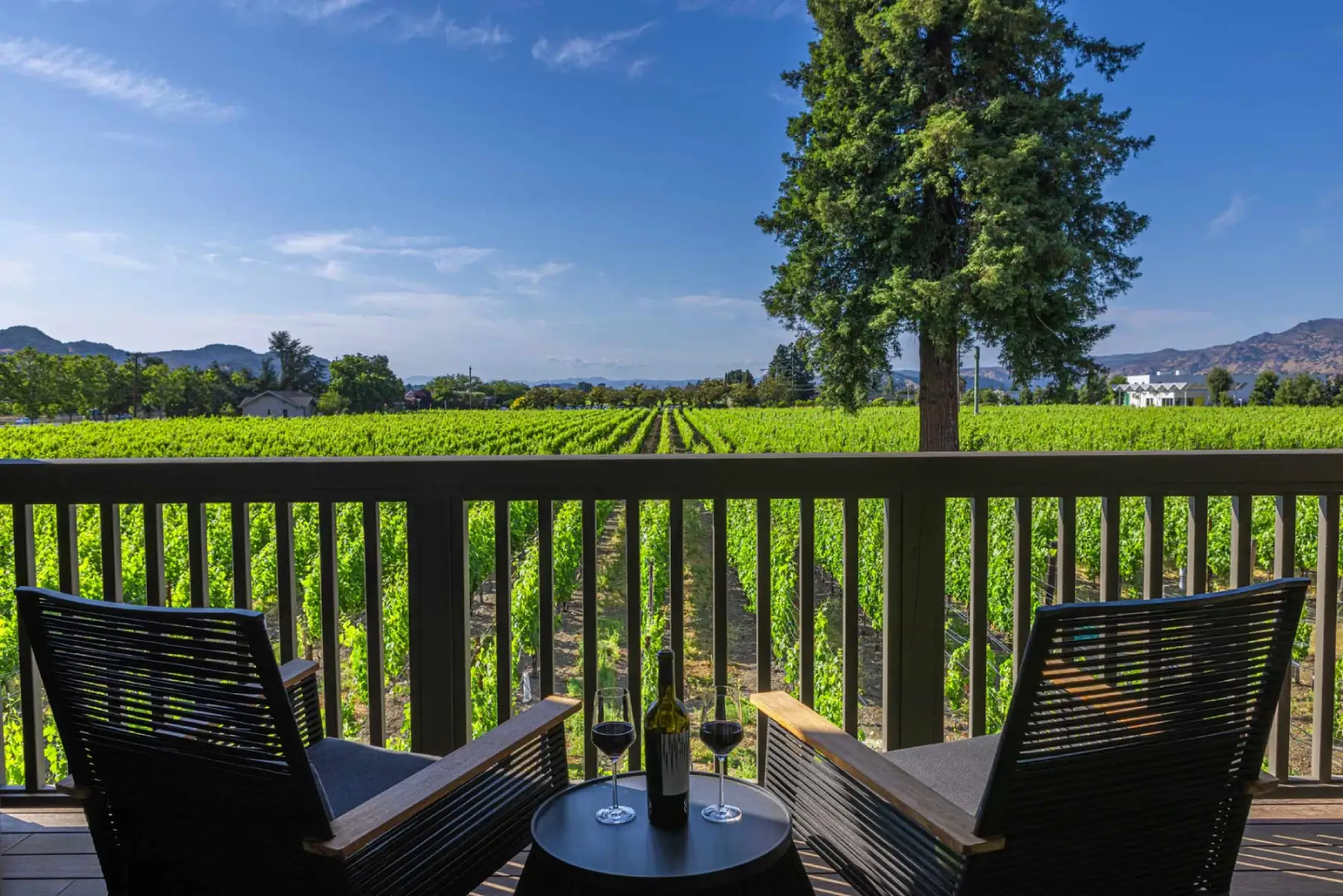 Two outdoor chairs and wine on a balcony with a stunning view of green napa valley vineyard rows.