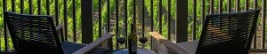 Two comfortable black chairs on a balcony overlooking lush greenery, with a bottle and glasses of wine on a small table.