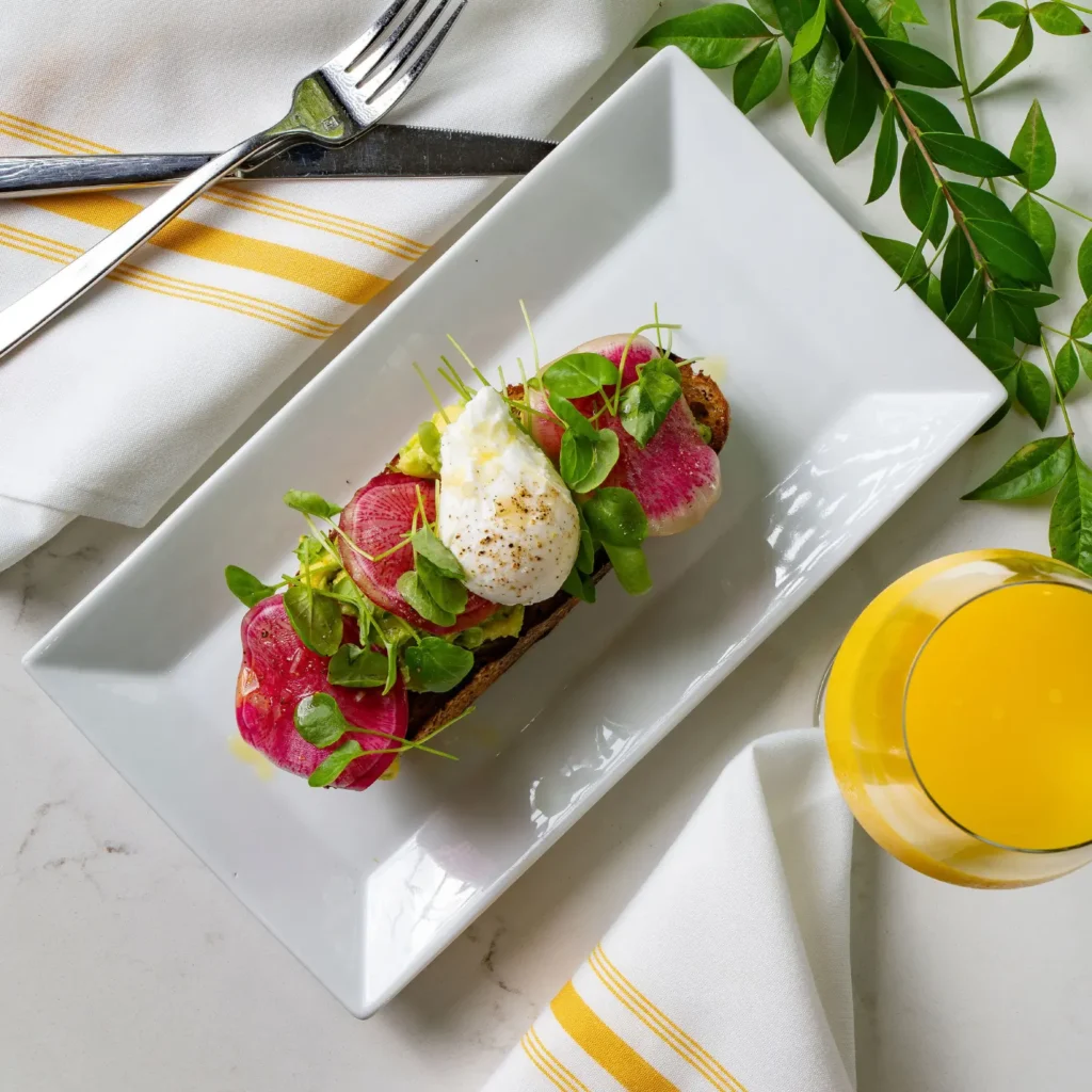 A top-down shot of avocado toast topped with a poached egg, sliced red radishes, and fresh microgreens, served on a rectangular white plate with a side of orange juice.