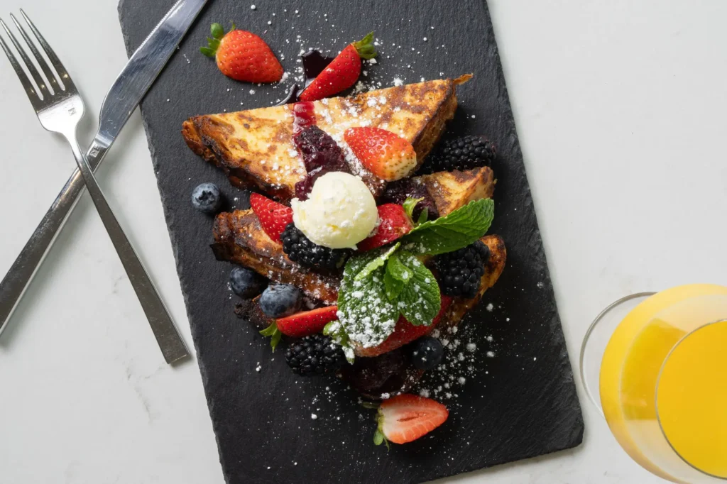 Brioche French Toast topped with a scoop of butter, fresh berries, mint, and powdered sugar on a black slate platter.