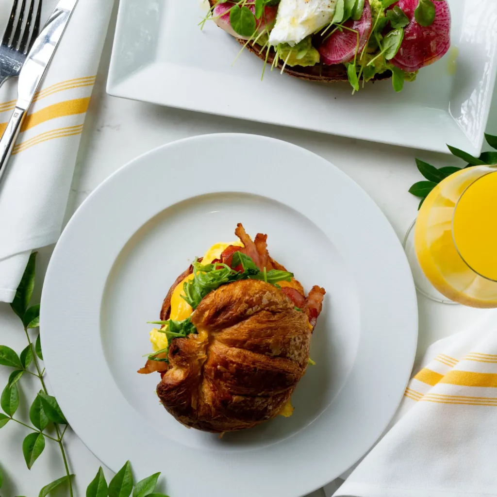 Overhead view of a bacon and egg croissant sandwich next to avocado toast topped with radish and a poached egg.
