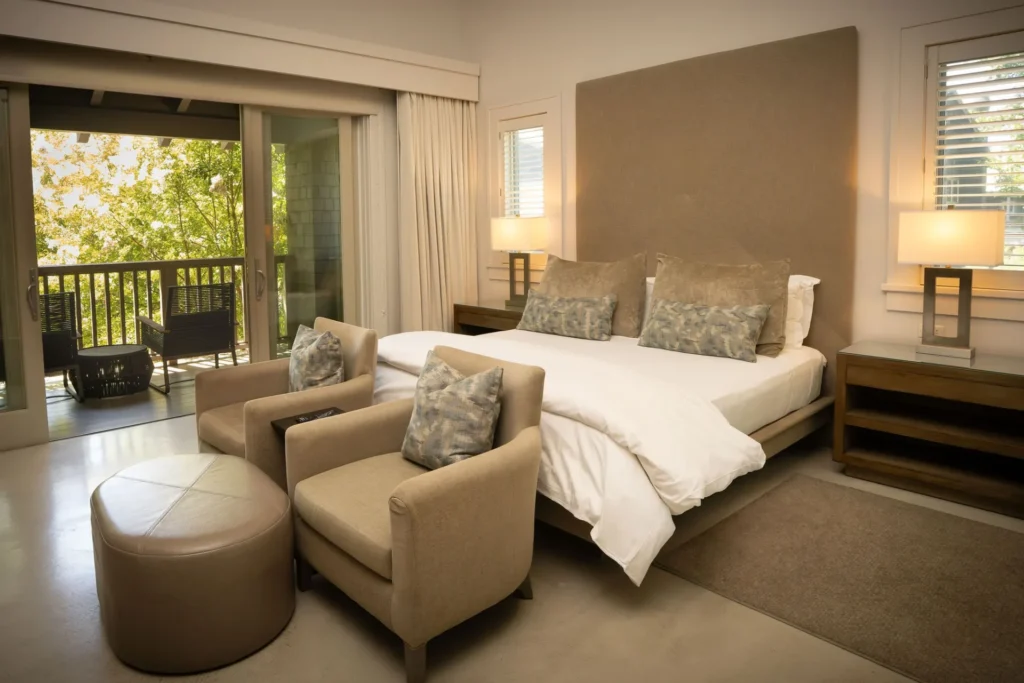 Modern hotel bedroom with king bed, beige armchairs, and balcony access