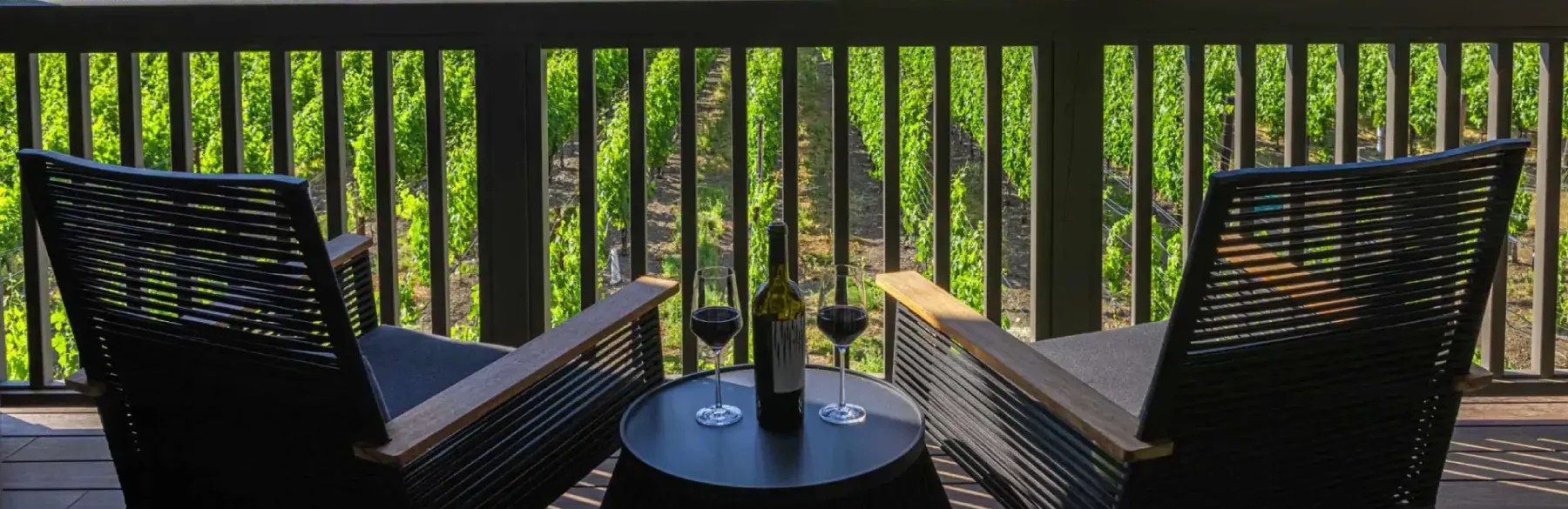 Two comfortable black chairs on a balcony overlooking lush greenery, with a bottle and glasses of wine on a small table.