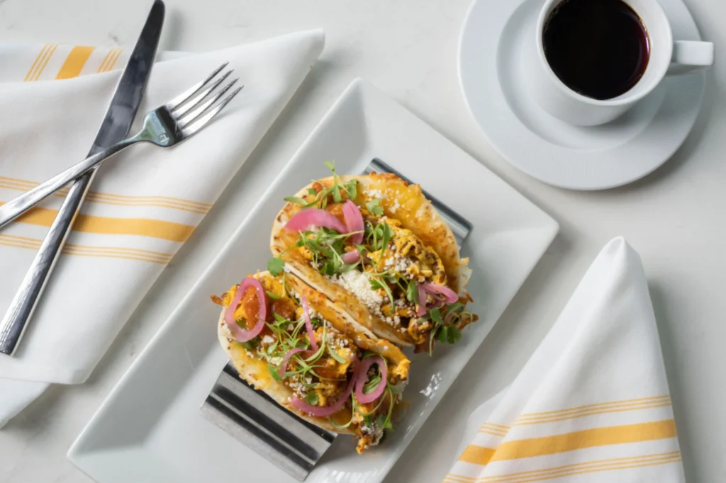 Two breakfast tacos garnished with pickled onions and cotija cheese served on a white platter with a cup of black coffee on the side.