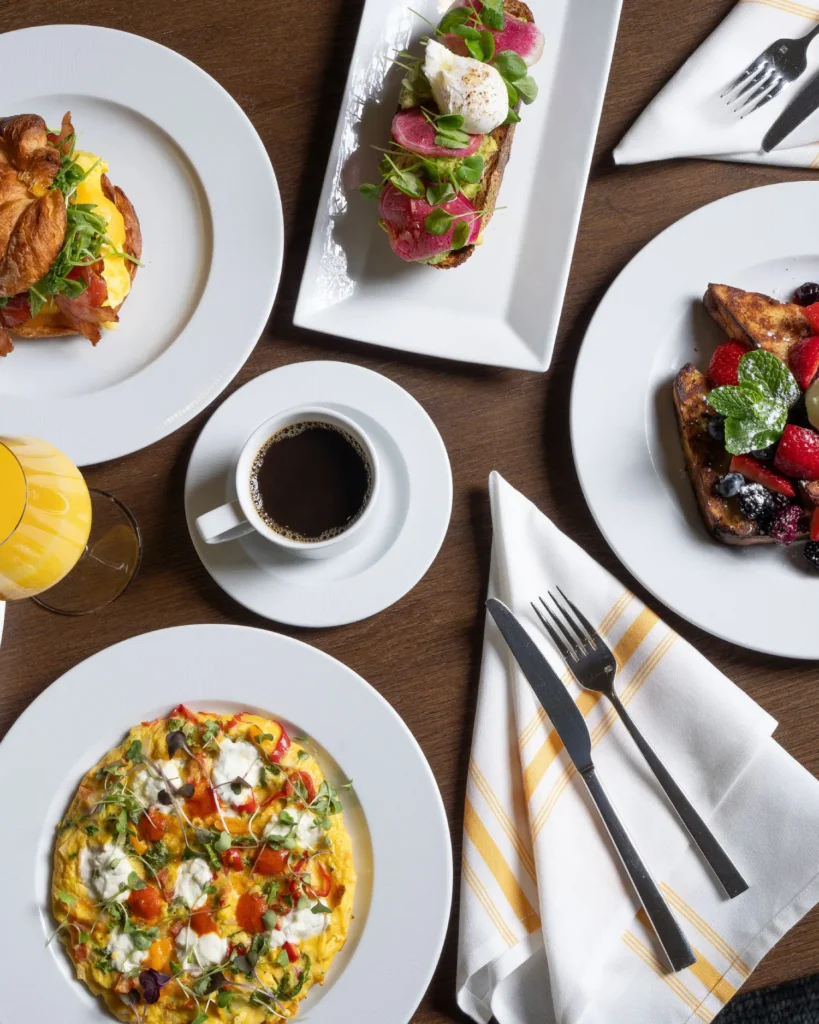 A top-down view of a gourmet breakfast spread on a dark wood table including a frittata with goat cheese, avocado toast with a poached egg, a croissant sandwich, berry-topped French toast, black coffee, and orange juice.