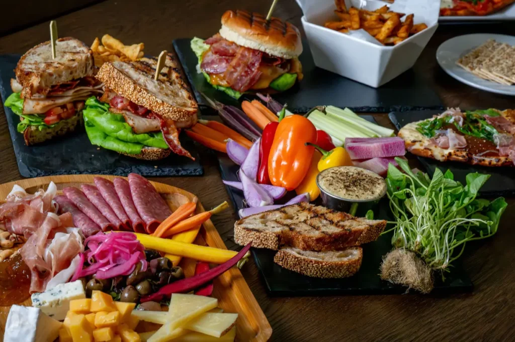 Assorted restaurant platters featuring sandwiches, charcuterie, and fresh vegetables on a wooden table.