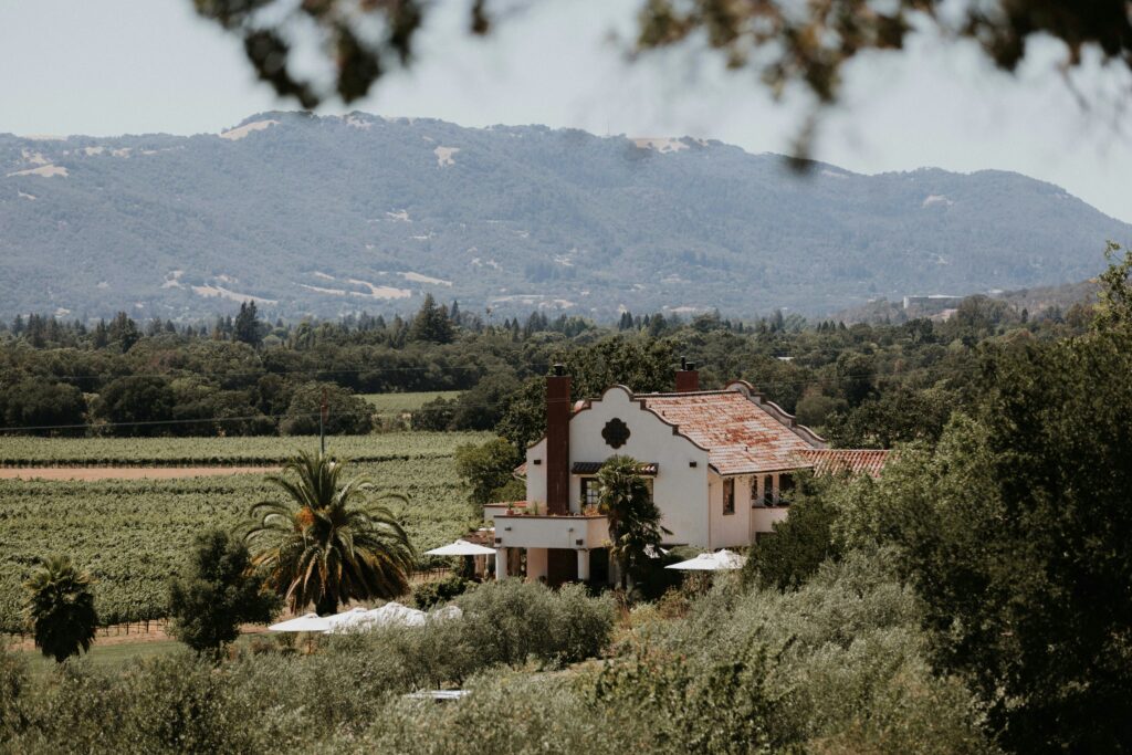 A scenic, high-angle view of a white Mediterranean-style winery estate with a terracotta roof, surrounded by lush green vineyard rows, palm trees, and rolling California mountains under a clear sky.