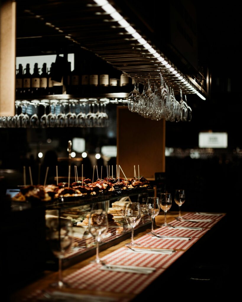A dimly lit, cozy restaurant bar counter displaying a variety of artisan tapas on toothpicks, with empty wine glasses set on a red-and-white checkered runner and more glasses hanging from an overhead rack.